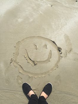 pexels-photo-698899-698899 A smiley face drawn in sand on a sunny beach, captured with shoes visible.